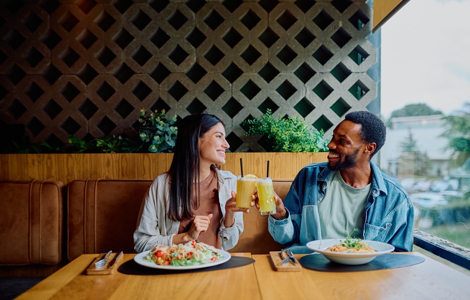 Casal felizes desfrutando de um almoço romântico em um restaurante, brindando com bebidas geladas e sorrindo um para o outro, com uma incrível vista ao fundo.