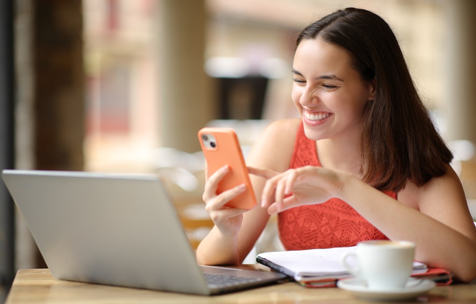Jovem mulher sorridente usando smartphone enquanto trabalha com laptop em ambiente de café, transmitindo felicidade e produtividade, sendo ideal para conteúdo sobre uso de tecnologia, trabalho remoto ou comunicação digital.