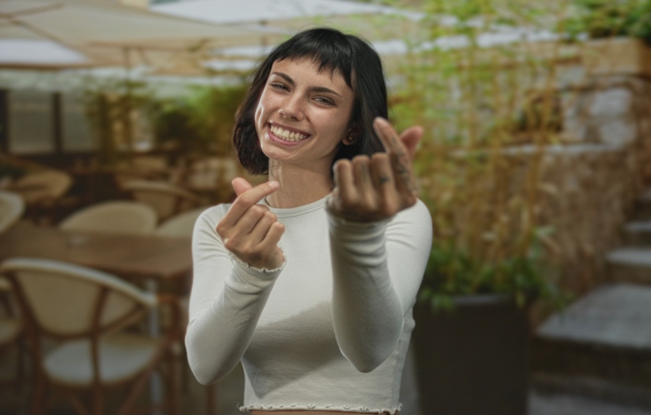 Jovem mulher sorrindo e fazendo gesto com as mãos, em ambiente ao ar livre com decoração de jardim, transmitindo alegria e descontração.