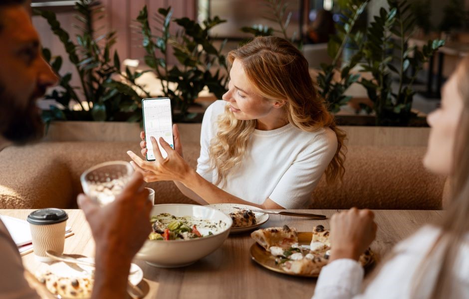 Homem sorridente, possivelmente um barista ou proprietário de cafeteria, usando um tablet para gerenciar pedidos ou promoções em um ambiente aconchegante de café, decorado com plantas e utensílios de cozinha. Ideal para conteúdo sobre gestão de cafeterias, empreendedorismo ou experiência em cafeterias.