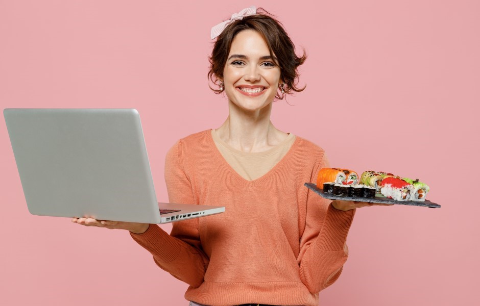Mulher sorridente com cabelo cacheado segurando um prato com sushi e usando um computador portátil, em fundo rosa, celebrando o consumo de sushi.