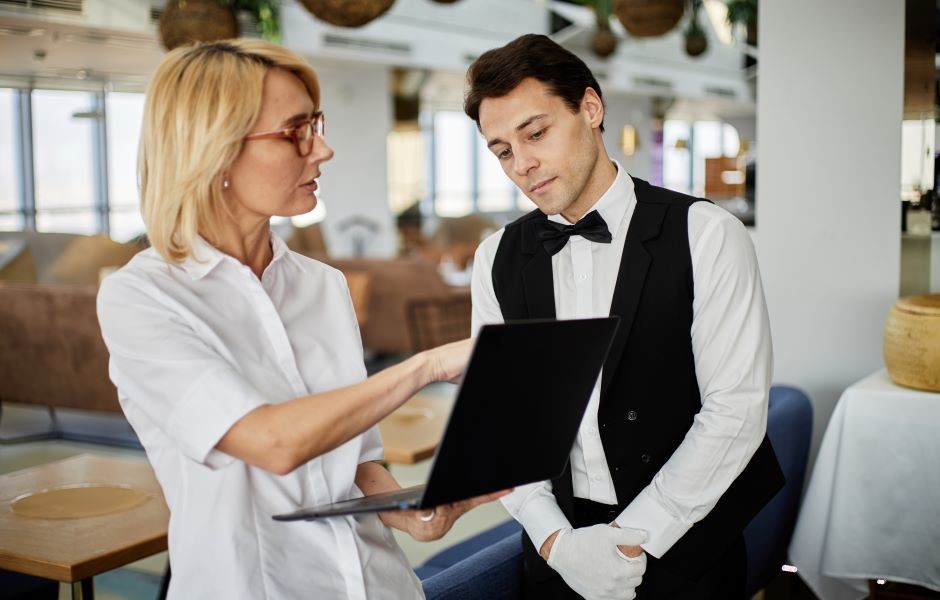 Homem vestindo uniforme de garçom com gravata borboleta, conversando com uma mulher com óculos e cabelo loiro em um restaurante durante o dia, enquanto verificam um menu digital.