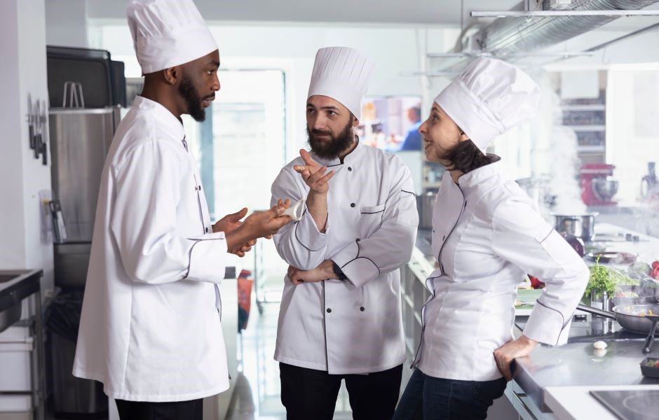 Chef de cozinha liderando uma equipe em discussão na cozinha de um restaurante, todos usando uniformes e chapéus de chef.