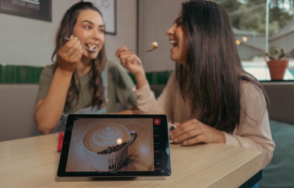 Duas amigas felizes desfrutando de um momento na cafeteria, com uma xícara de café com latte art e um tablet na mesa, em um ambiente acolhedor.