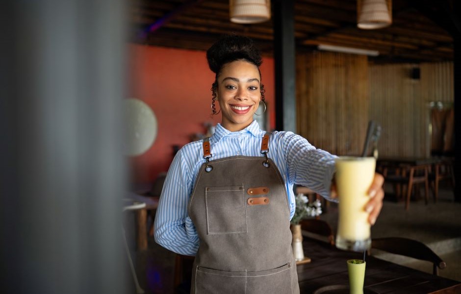 Mulher sorridente em um restaurante, usando avental e oferecendo uma bebida, representando serviço de atendimento ao cliente em bares ou restaurantes.