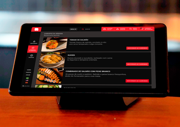 Tela de um aparelho de mesa mostrando um cardápio digital de restaurante japonês com opções de sushi e salmão, em ambiente de restaurante.