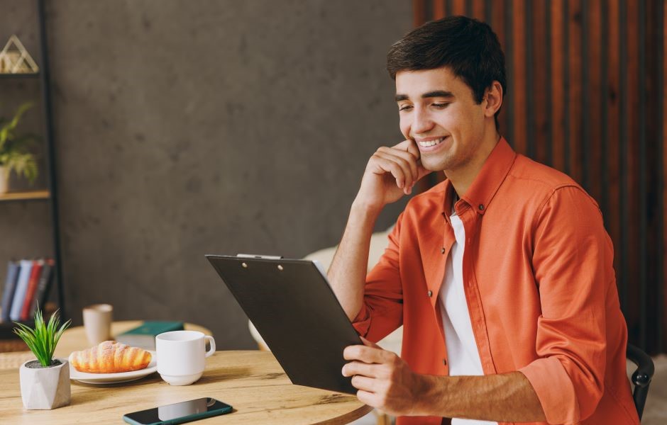 Homem sorrindo usando um tablet, sentado à mesa com uma xícara, prato com croissants e um telefone na mesa, ambiente de escritório ou home office.