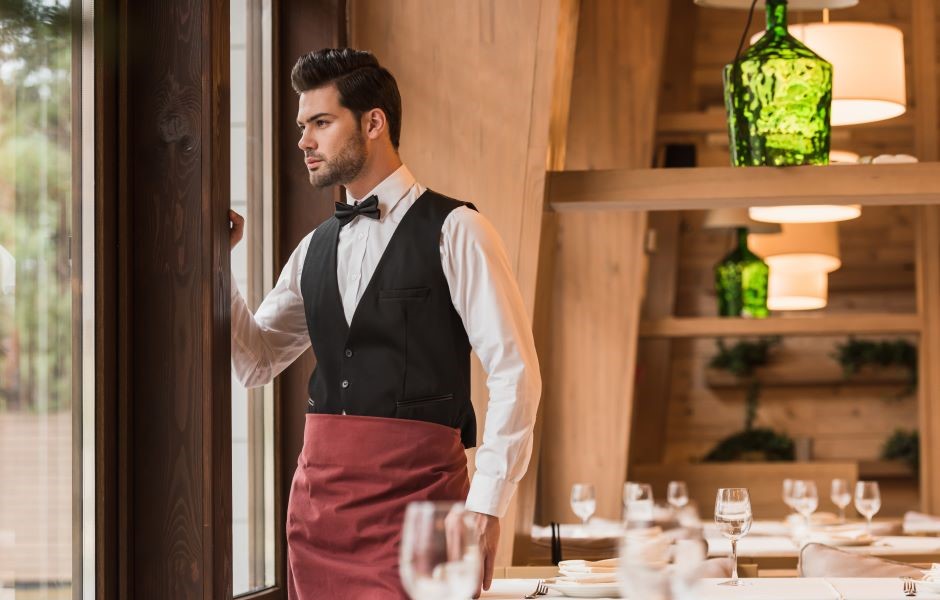 Homem elegante em uniforme de garçom, usando terno com gravata borboleta, em restaurante sofisticado com mesas bem decoradas e iluminação aconchegante.