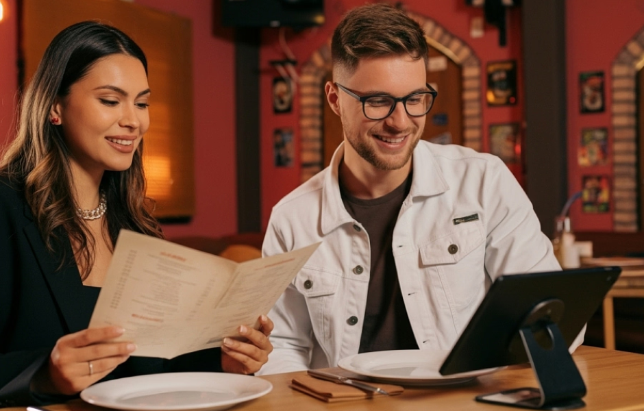 Casal jovem sorridente em restaurante moderno, com a mulher segurando o cardápio e o homem usando tablet, ambiente aconchegante com decoração colorida.