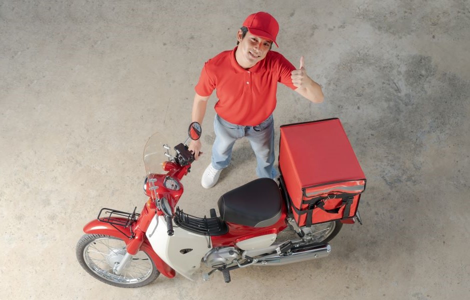 Homem sorridente vestindo uniforme de entregador de pizza com camiseta vermelha e boné, ao lado de uma moto de entrega vermelha, preparado para fazer entregas.