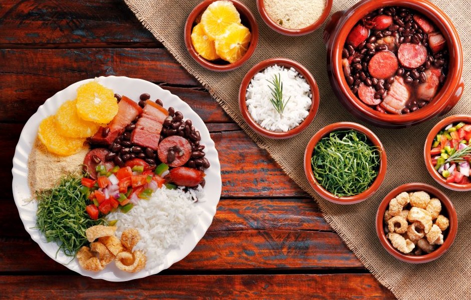 Prato de feijoada com arroz, couve, farofa, laranja e acompanhamentos tradicionais em tigelas de barro, servido em mesa de madeira rústica, destacando-se na culinária brasileira.