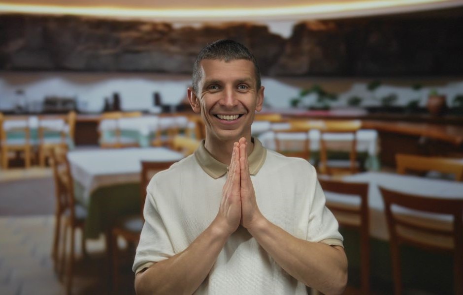 Homem sorridente com as mãos juntas em oração, em restaurante aconchegante, representando gratidão e paz interior, promovendo bem-estar e espiritualidade.
